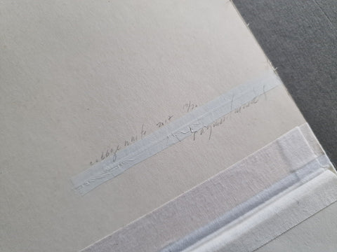 A close-up of a textured off-white paper sheet with faint handwritten text on a strip of translucent tape, placed on a dark gray surface.