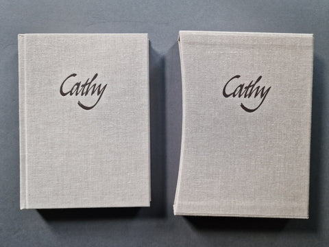 The linen book cover and its matching slipcase side by side on a dark gray background, both featuring the name 'Cathy' printed in black cursive script on the front cover.
