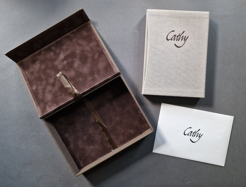 A limited edition box set featuring an open dark brown velvet-lined presentation box with a ribbon, a matching textured cream book titled Cathy in a slipcase, and a white card, all personalized with the name 'Cathy' in black script, arranged on a dark gray surface.