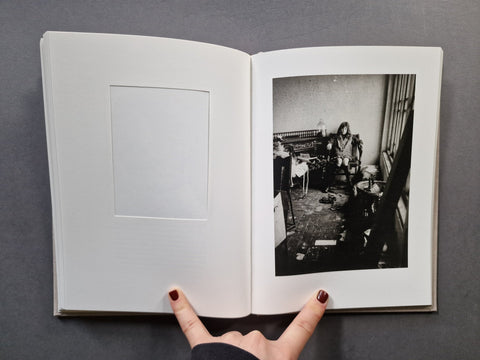 An open hardcover book: The left page has a blank white mat and inset frame, while the right page shows a black-and-white photograph of a cluttered interior scene and a girl.