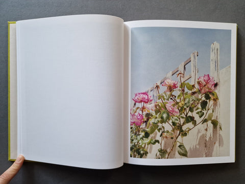 An open book showing a blank white left page and a printed photograph on the right page of pink flowers growing along a white fence under a pale blue sky.