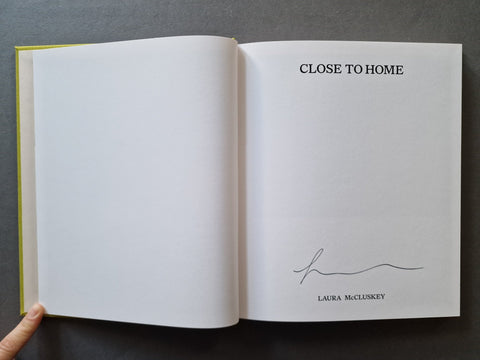 An open book, showing a blank white left page and a title page on the right with the text 'CLOSE TO HOME,' 'LAURA McCLUSKEY,' and a handwritten signature.