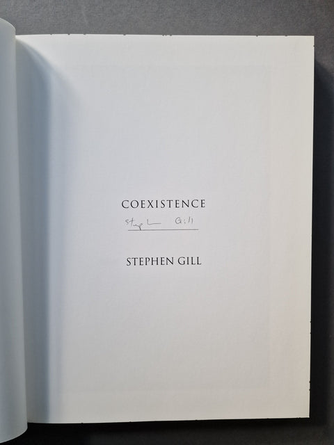 An open book showing a white interior page with the title 'COEXISTENCE', a handwritten signature, and the author name 'STEPHEN GILL' printed in black serif text.