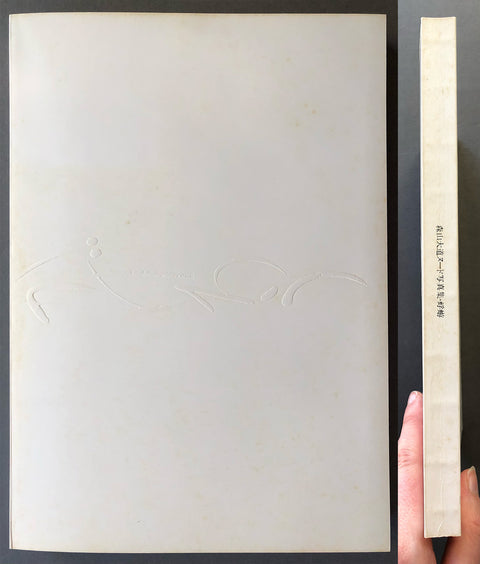 A hand holds a tall, narrow book or booklet standing beside a large off-white rectangular panel or cover. The main visible product is the tall booklet with a cream spine and black vertical text, while the large panel behind it is also off-white with a faint embossed abstract mark.