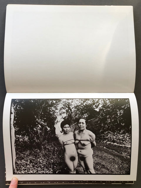 An open book or photo album showing a black-and-white printed photograph on the lower page of two nude people standing outdoors among trees, with a white page blank on the upper page.