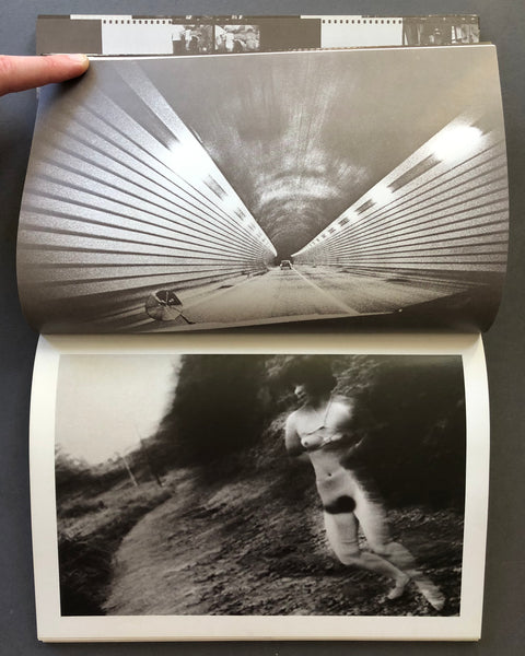 An open black-and-white art book or photo magazine being held by a hand, showing two printed photographs: a large tunnel scene on the top page and a darker outdoor figure scene on the bottom page.