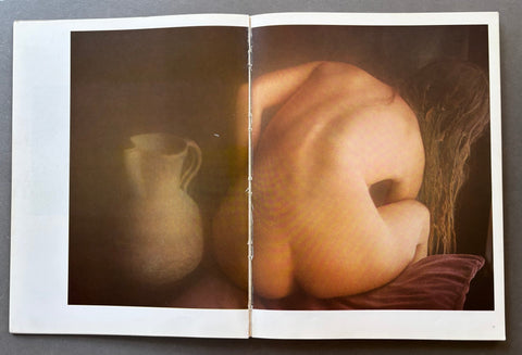 An open art book or magazine spread, showing a photographic print of a nude figure from behind next to a vase, photographed against a dark gray background.