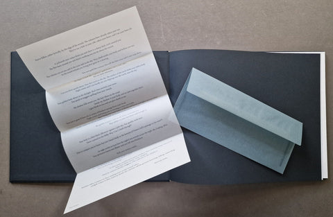 An open dark book or booklet laid on a neutral surface, with a folded white paper insert spread open on the left and a folded light blue envelope or card on the right.