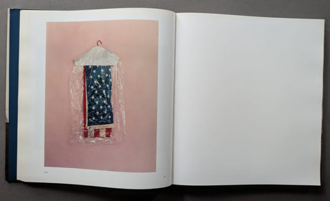 An open book showing a photograph of a small American flag hanging in a clear plastic bag against a pink background, with the right page blank.