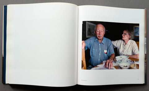 An open book spread open on a dark surface, with a printed photograph on the right page showing an older man and woman seated at a table.