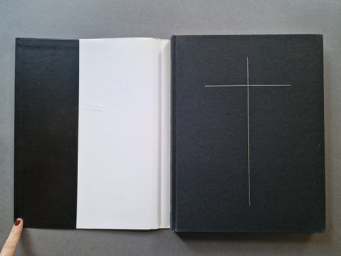 An open black book. The left page is white, and the right cover is black with a thin white cross design.