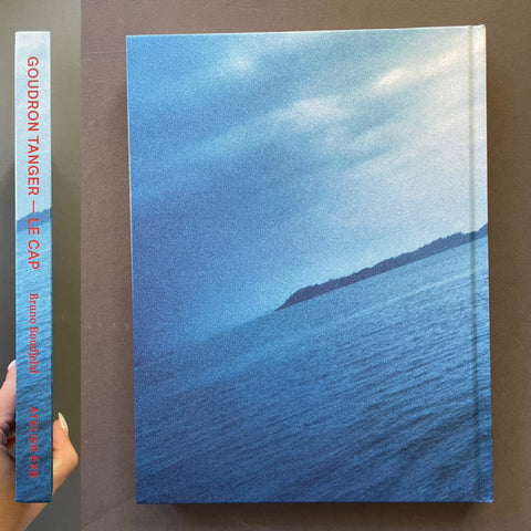 A hardcover book titled 'Goudron Tanger - Le Cap' by Bruno Boudjelal, featuring a blue-toned seascape cover and spine, photographed against a brown background with a hand holding the spine on the left.