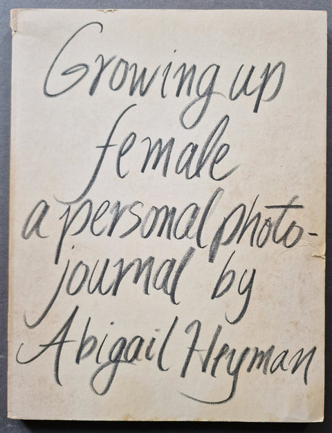 A vintage photobook cover with cream-colored cover and handwritten black ink text reading 'Growing up female a personal photo journal by Abigail Heyman'.