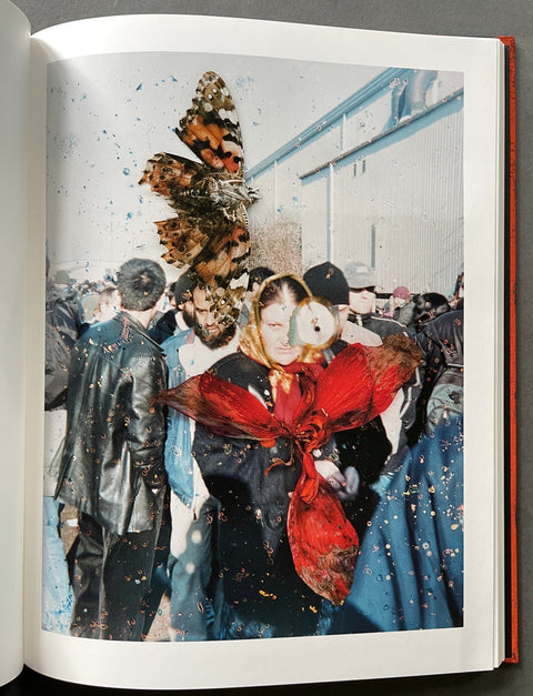 An open book showing a printed artwork or photo page with a surreal scene of people, large butterflies, and a red flower-like form, photographed against a dark gray background.