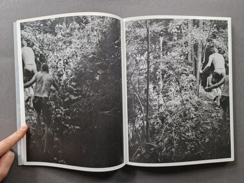 An open black-and-white art book or photo print showing two photographic panels of people in a dense forest, held open by a hand against a dark gray background.