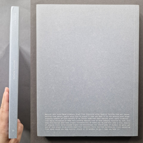 A hardcover book or journal shown from the back and spine, held by a hand against a dark gray background. The cover is a textured light gray with small white text at the bottom and vertical spine text.