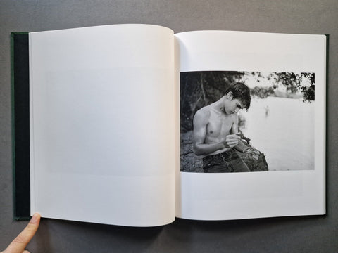 An open hardcover book. The left page is blank white, and the right page shows a black-and-white photograph of a shirtless man sitting outdoors near water.