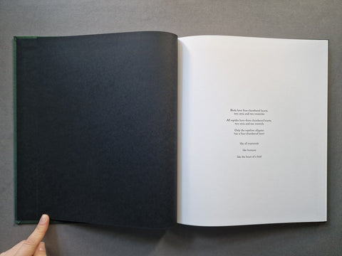 An open book lying on a dark gray surface, with a black cover on the left page and a white interior page with small black text on the right. A hand is visible holding the left edge of the book.