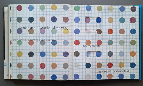 An open book showing two pages covered in a colorful pattern of paper circles on white graph paper, with printed text and a small cutout window on the right page.
