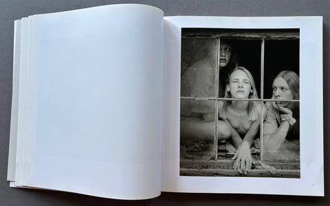 An open art book or photo journal laid flat on a dark surface, showing a large black-and-white photographic print on the right page with three seated figures behind a barred window, and an empty white left page.