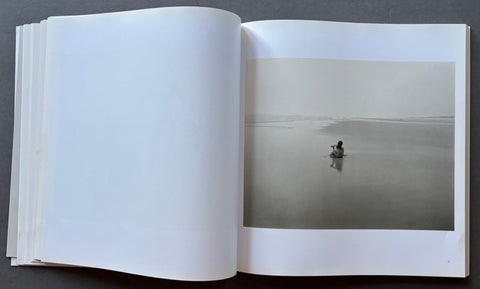 An open book or photo journal laid flat on a dark gray surface, showing a large white left page and an open right page with a minimalist black-and-white landscape photograph of a small figure in a misty flat landscape.
