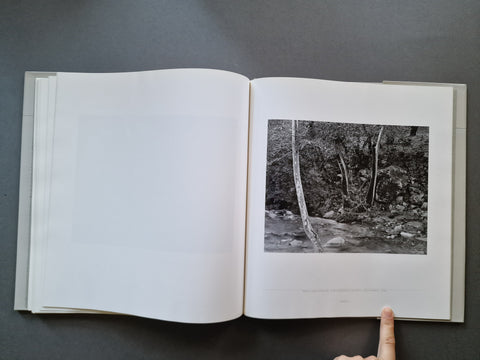 An open hardcover book, showing a black-and-white landscape photograph printed on the right page. A person's finger is pointing at the photograph.