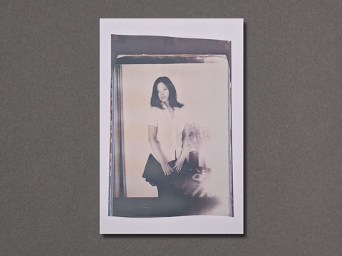 A vertical photographic art print with a white border. The photograph depicts a woman standing dressed in a white shirt and black skirt