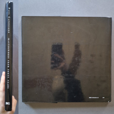 A square dark book cover with a reflective surface showing a person taking a mirror selfie, placed on a gray surface next to a tall black spine labeled 'PAUL Caponigro' and 'PWG'.