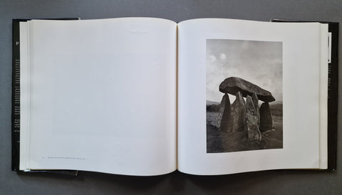 An open book showing a black-and-white photograph of a stone megalithic structure on the right page, with blank white pages on the left, photographed from above on a dark gray surface.