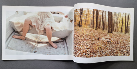 An open art photo book, showing two printed photographs side by side. The left page features a photo of bare legs seated in a bathtub, and the right page shows a forest scene with fallen leaves and a person lying on the ground.
