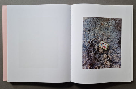An open book, showing a mostly blank white left page and a printed photograph on the right page of a small colorful rectangular item placed on rough gray-brown textured ground.