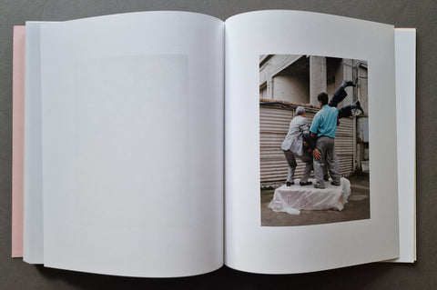 An open book, with mostly blank white pages on the left and a printed photograph on the right showing two people standing outdoors near a corrugated metal wall.