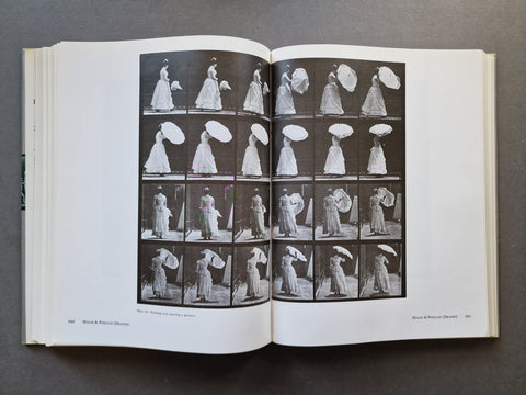 An open vintage book showing a black-and-white photographic plate sequence of a woman in a long dress holding a parasol, photographed from above on a dark gray surface.