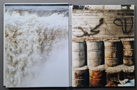 An open art book spread showing two photographic scenes side by side: a high-contrast image of rushing white water on the left, and a gritty scene with stacked metal barrels and graffiti on the right.