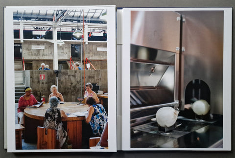 A square art book spread showing two photographic scenes side by side: on the left, an interior scene with several people seated around a round table in a concrete-walled hall; on the right, a close-up of a stainless steel industrial machine with white spherical objects in front of a dark opening.