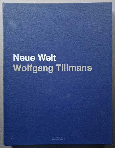 A hardcover book titled 'Neue Welt Wolfgang Tillmans' in white text on a textured dark blue cover, photographed flat against a neutral gray background.