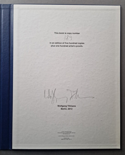 The interior title page of a hardcover book, showing a white page with printed text, a handwritten copy number 189, a signature, and Wolfgang Tillmans' name and Berlin, 2012 location. The book's dark blue cloth binding is visible along the left edge against a dark background.