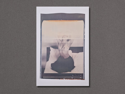 A vertical photographic art print with a white border. The photograph depicts a person sitting on a bed in a monochrome, slightly faded style with a warm beige tint at the top.