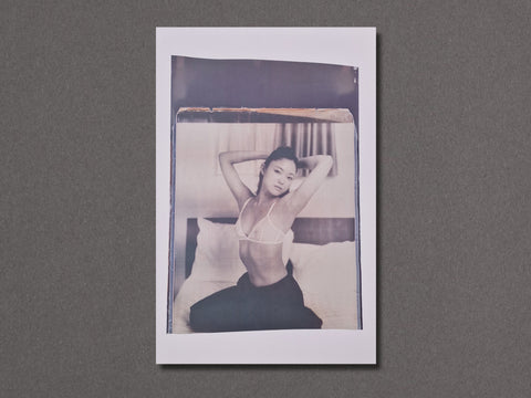 A vertical photographic art print with a white border. The photograph depicts a person sitting on a bed in a monochrome, slightly faded style with a warm beige tint at the top. Close up shot