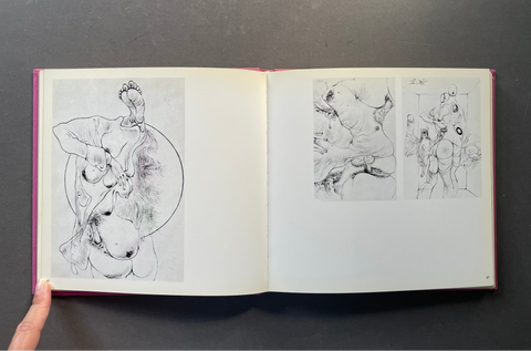 An open art book or sketchbook on a dark gray surface, showing black line drawings and sketches printed on white pages. A hand is visible holding the lower left edge of the book.