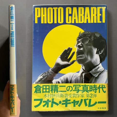 A hardcover book titled 'Photo Cabaret' shown standing upright next to its spine, with a hand holding the spine for scale. The cover features a large yellow sun graphic, bold yellow title text, and a black-and-white portrait of a man wearing glasses against a blue background.