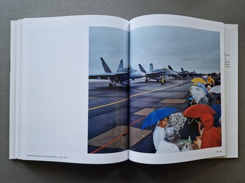 An open book showing a large color photograph of fighter jets on an aircraft carrier deck, with onlookers holding umbrellas in the rain in the lower right of the print.