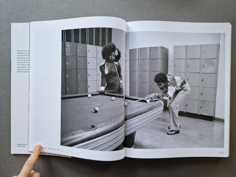 An open book showing a black-and-white archival photograph of two people playing pool in a locker room. background.
