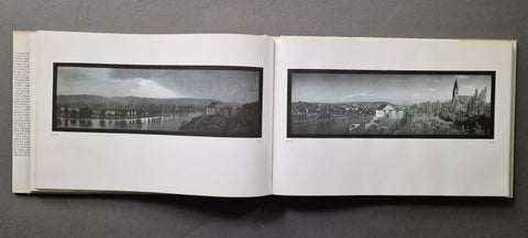An open vintage book spread on a dark gray surface, showing two black-and-white panoramic landscape photographs printed on white pages.