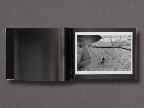 An open black-and-white photo book or art print set displayed flat against a dark gray background. The left page is mostly dark, while the right page shows a black-and-white photograph of a person in profile and a small bird on a cobblestone surface.