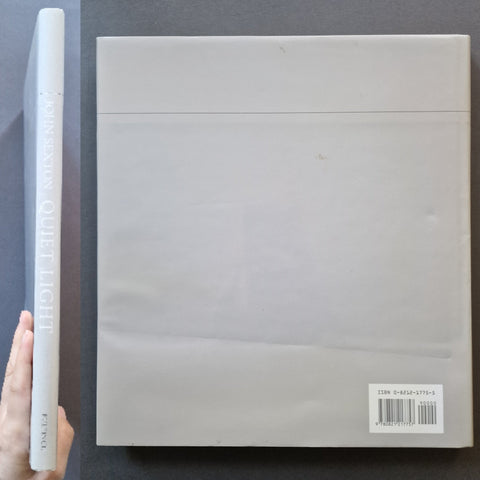 The back cover of the book 'John Sexton Quiet Light', shown next to its spine. A hand is holding the spine on the left to show scale against a dark gray background.