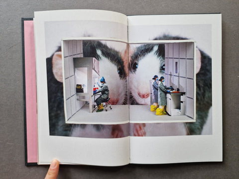 An open book - The pages show a surreal printed image: a close-up of two rats behind a window-like inset, with a small clinical room scene inside.