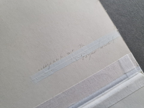 A close-up of a textured off-white paper sheet with faint handwritten text on a strip of translucent tape, placed on a dark gray surface.