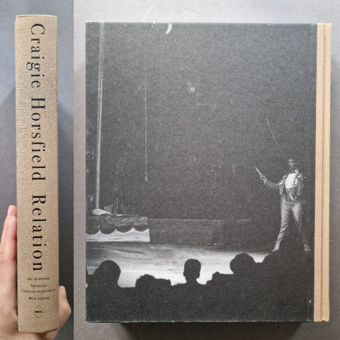 A hardcover book titled 'Craigie Horsfield Relation' is shown standing upright next to a hand holding the spine. The cover features a black-and-white photograph of a performer on stage, with the title on a beige textured spine.
