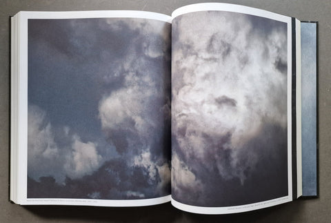 An open hardcover book displaying a large, high-contrast photograph of dramatic storm clouds across two pages, set against a dark textured background.